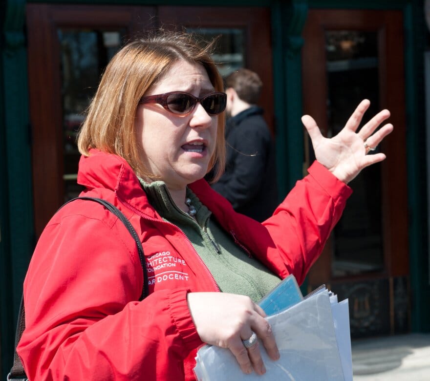 Tour guide in a red jacket in front of a building, gesturing with their left arm