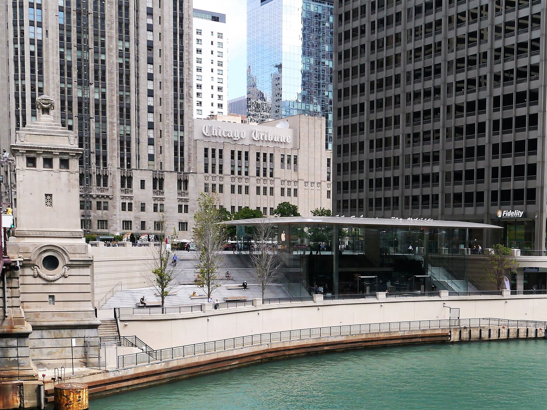 Image of a flat roof glass building on river front in the foregroundwith tall black and tan buildings in the background rising pout of frame.