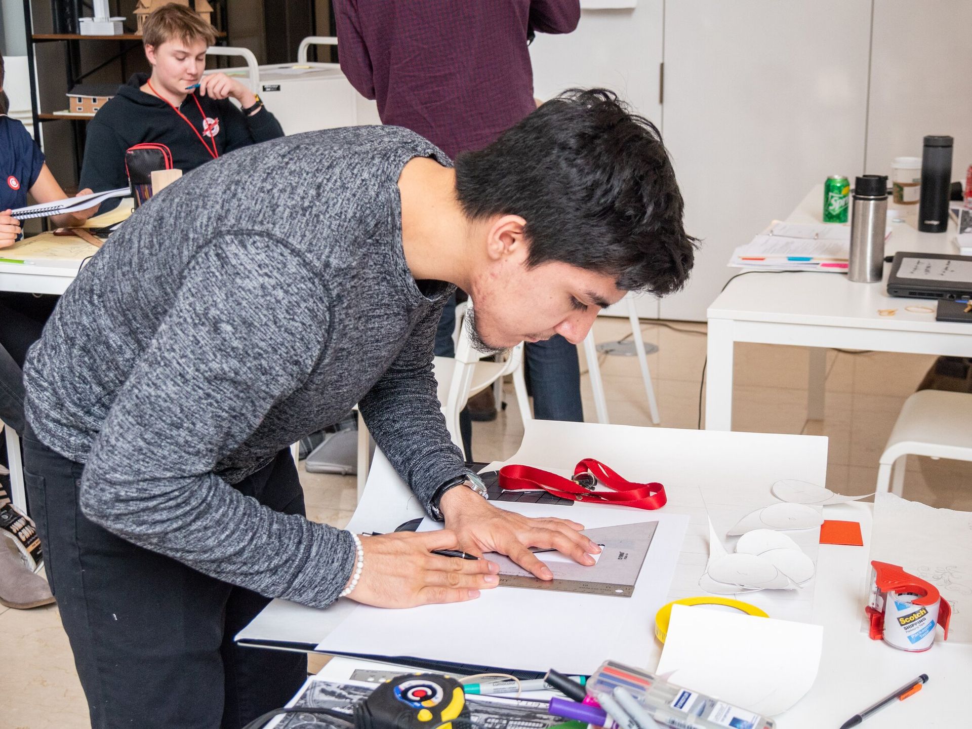 Man with dark hair in a gray shirt bending over a table and using a ruler to draw. 