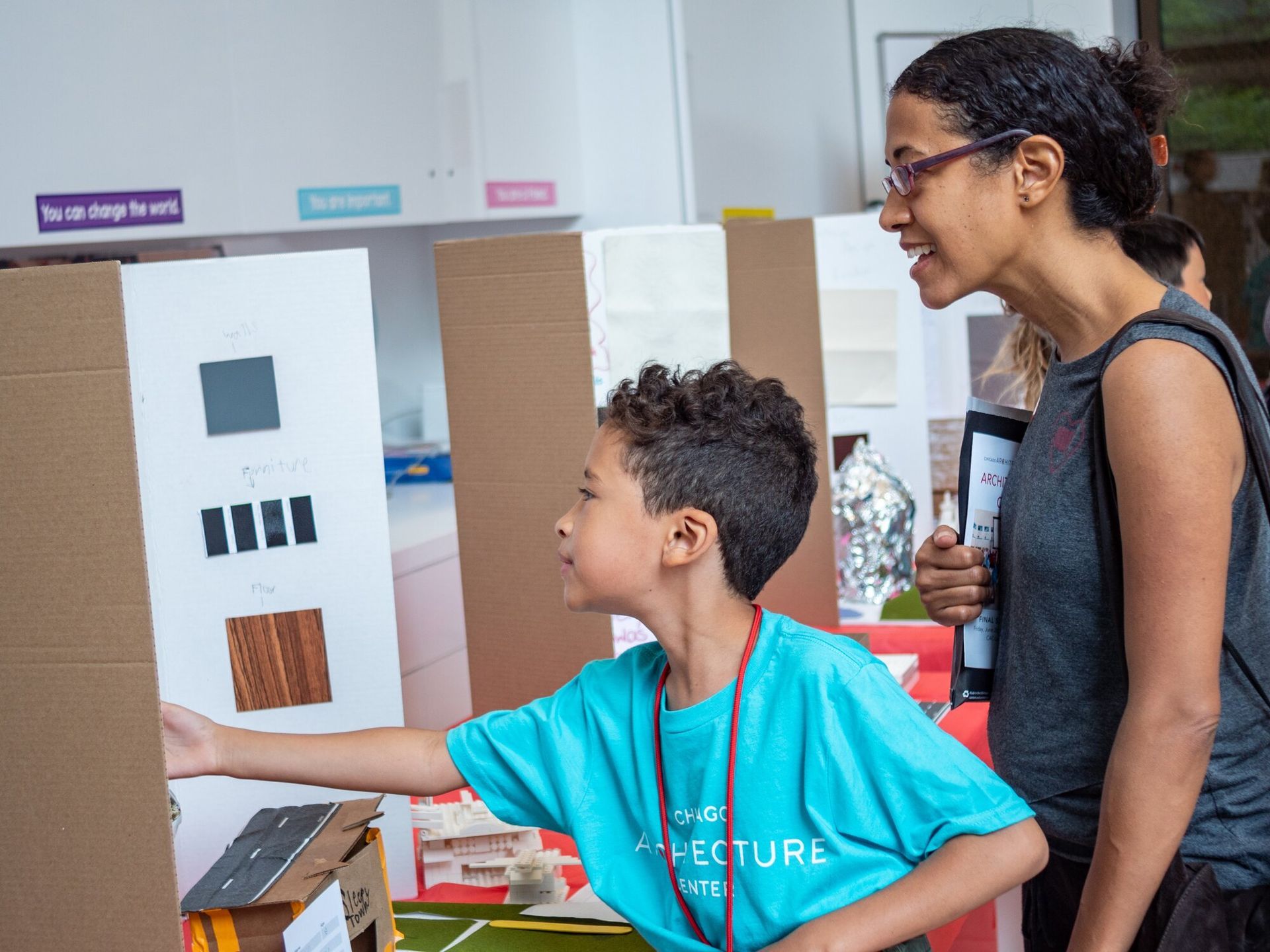 Young boy in a teal shirt pointing to a display next to a woman in glasses with dark hair.