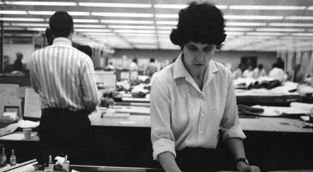 Black and white image of a woman with dark hair wearing a button up blouse working at a table and looking down.