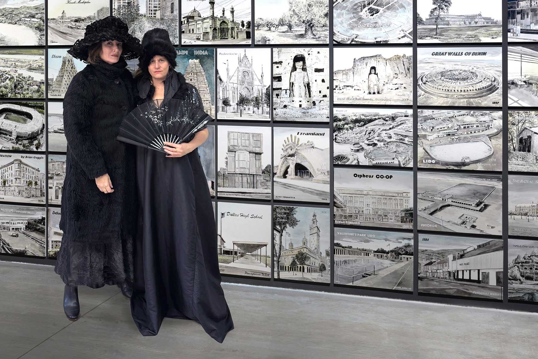 Two women dressed in black costumes standing in front of a wall of black and white photos.