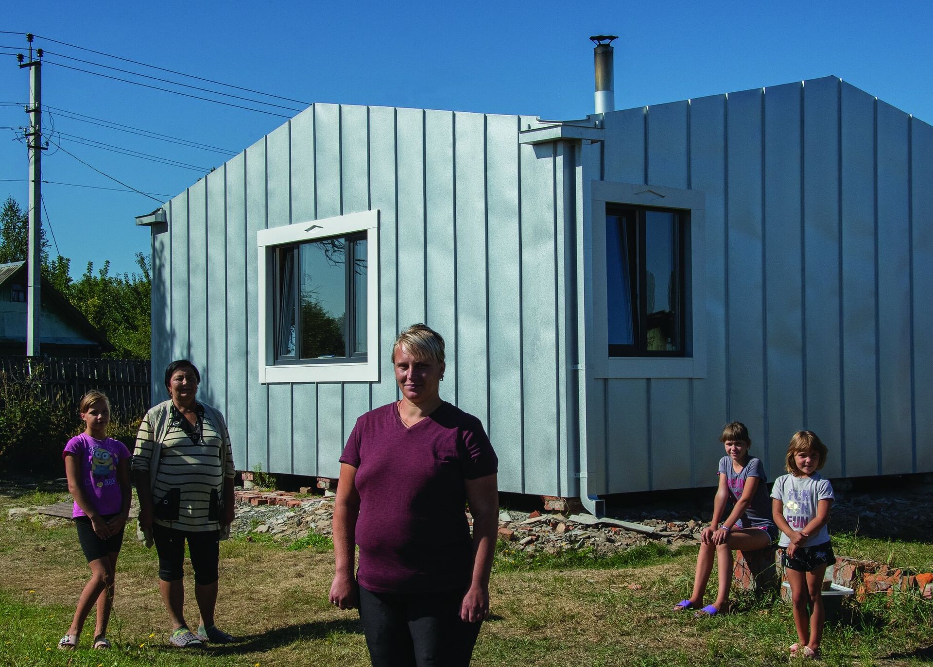 Image of a woman with short blonde hair standing in the foreground with two children to her right and a woman and child to her left. They all stand in front of a metal one story building.