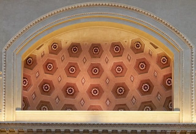 Ornate arched ceiling with squares carved into it above a large window framed by columns and lit with a red light