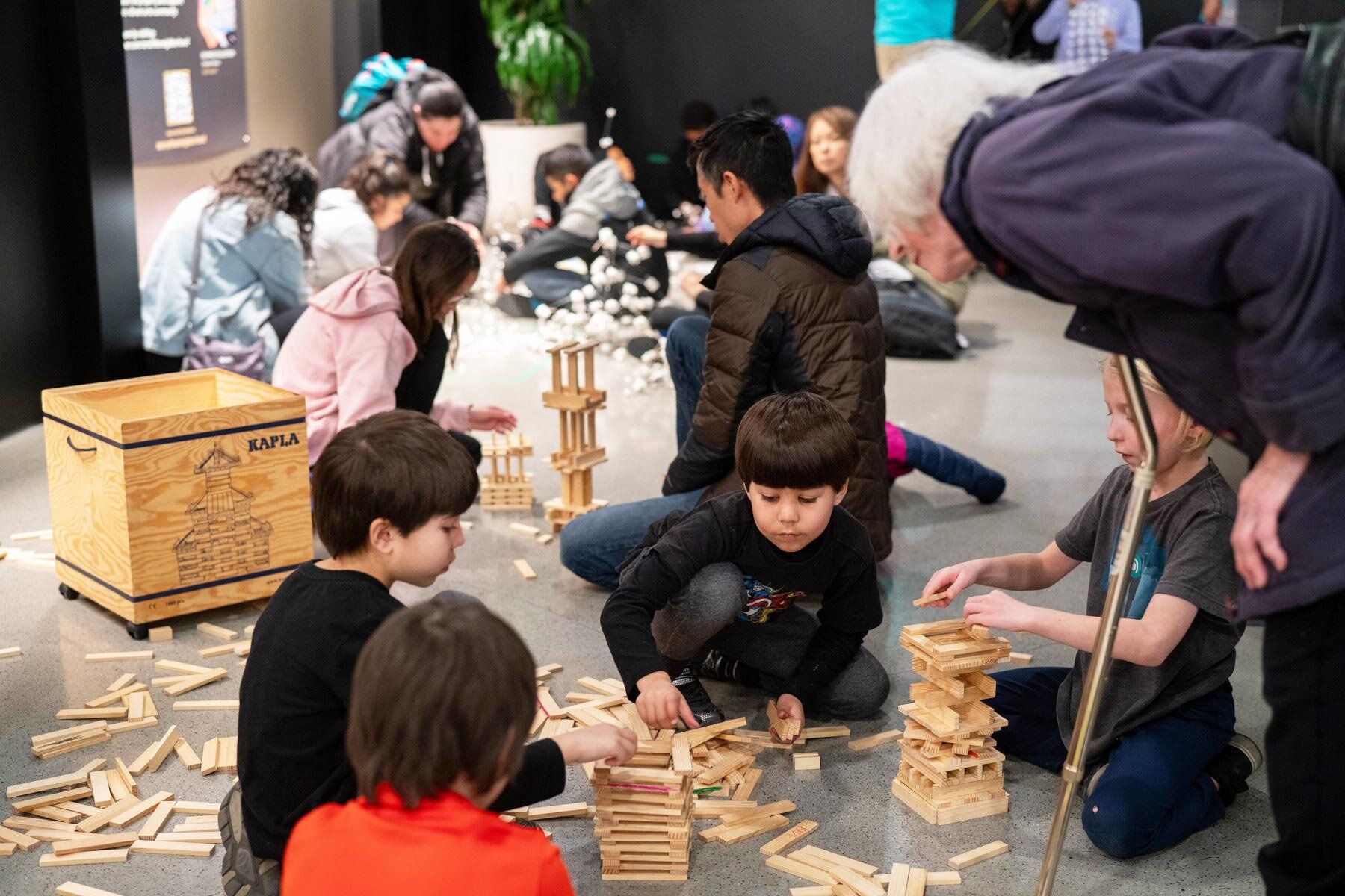 Children building wooden bricks on the floor while an elderly lady looks on