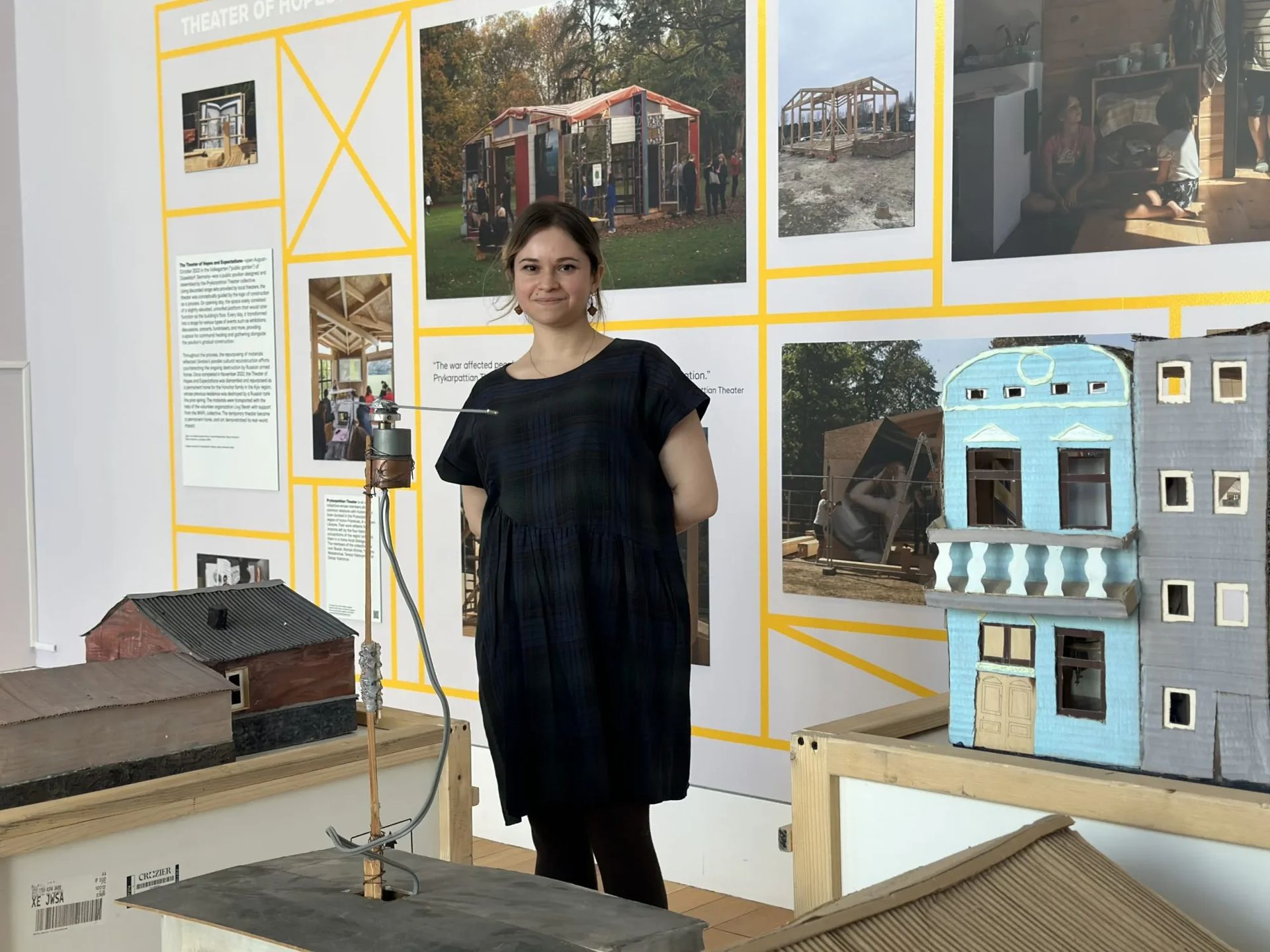 Image of a woman in a black short sleeve dress standing in an exhibition around wall panels covered in text and miniature models
