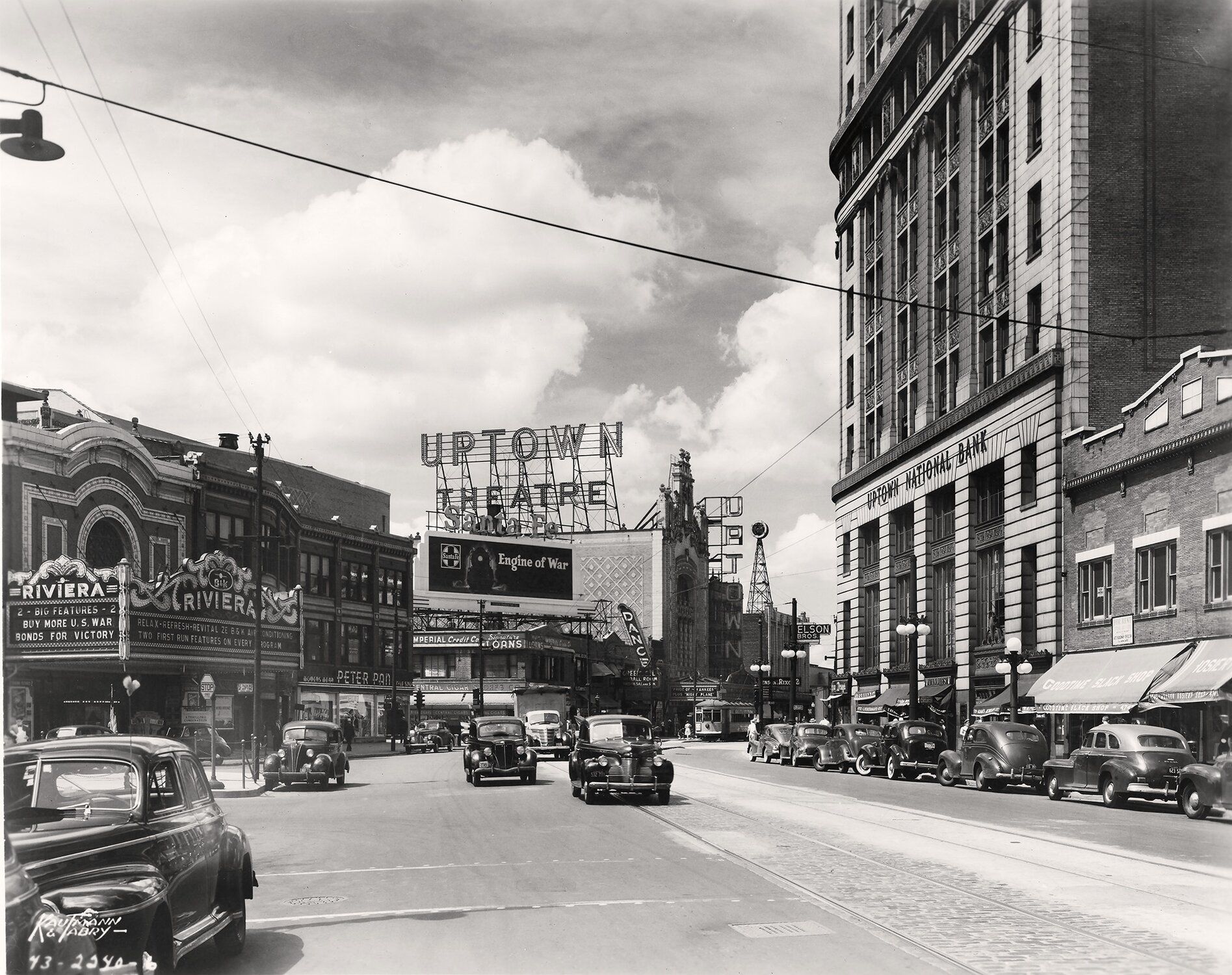 Black and white photo of a street with vintage cars and buildings on either side of the street. A sign in the middle reads "Uptown Theatre"