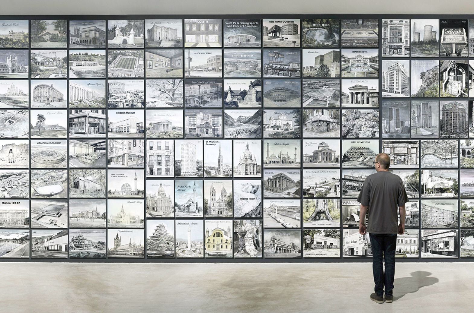 Man standing and facing away from the viewer looking at a wall of illustrations of different locations.