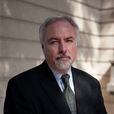 Headshot of a caucasian man with a beard in a black suit jacket standing against a wall