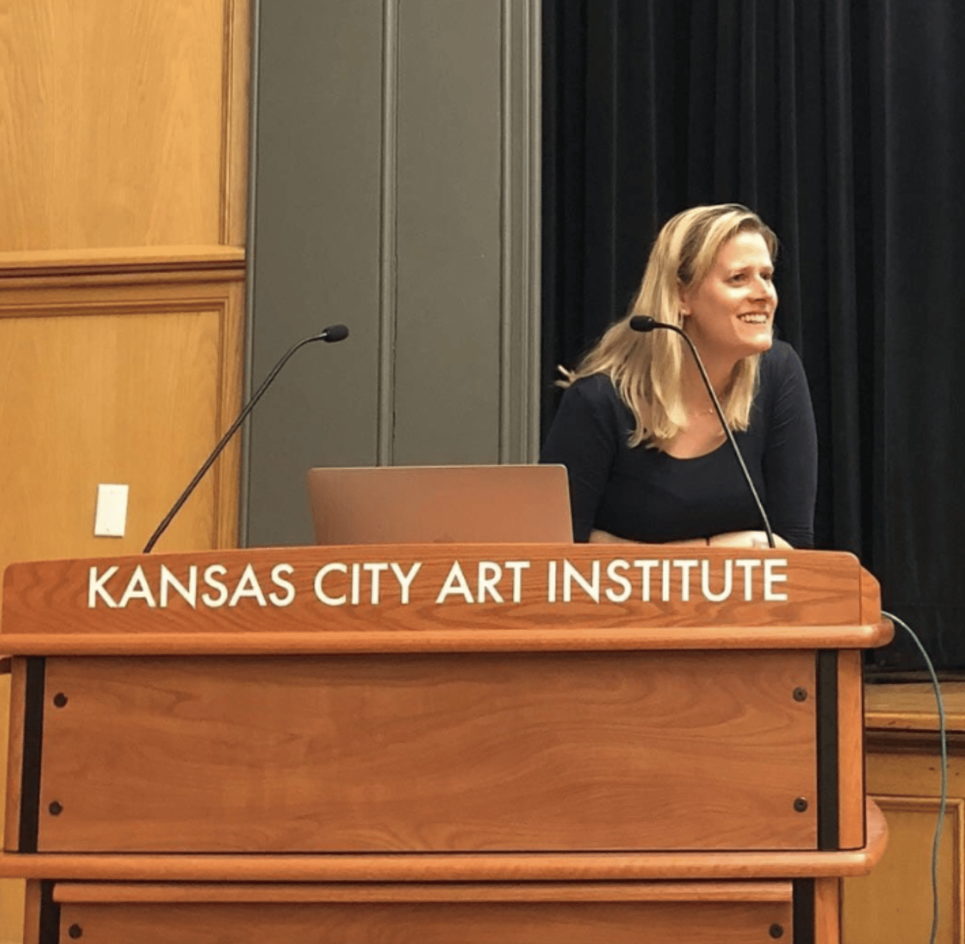 Women with blonde hair and a black top, looking away from the camera, standing in front of a podium