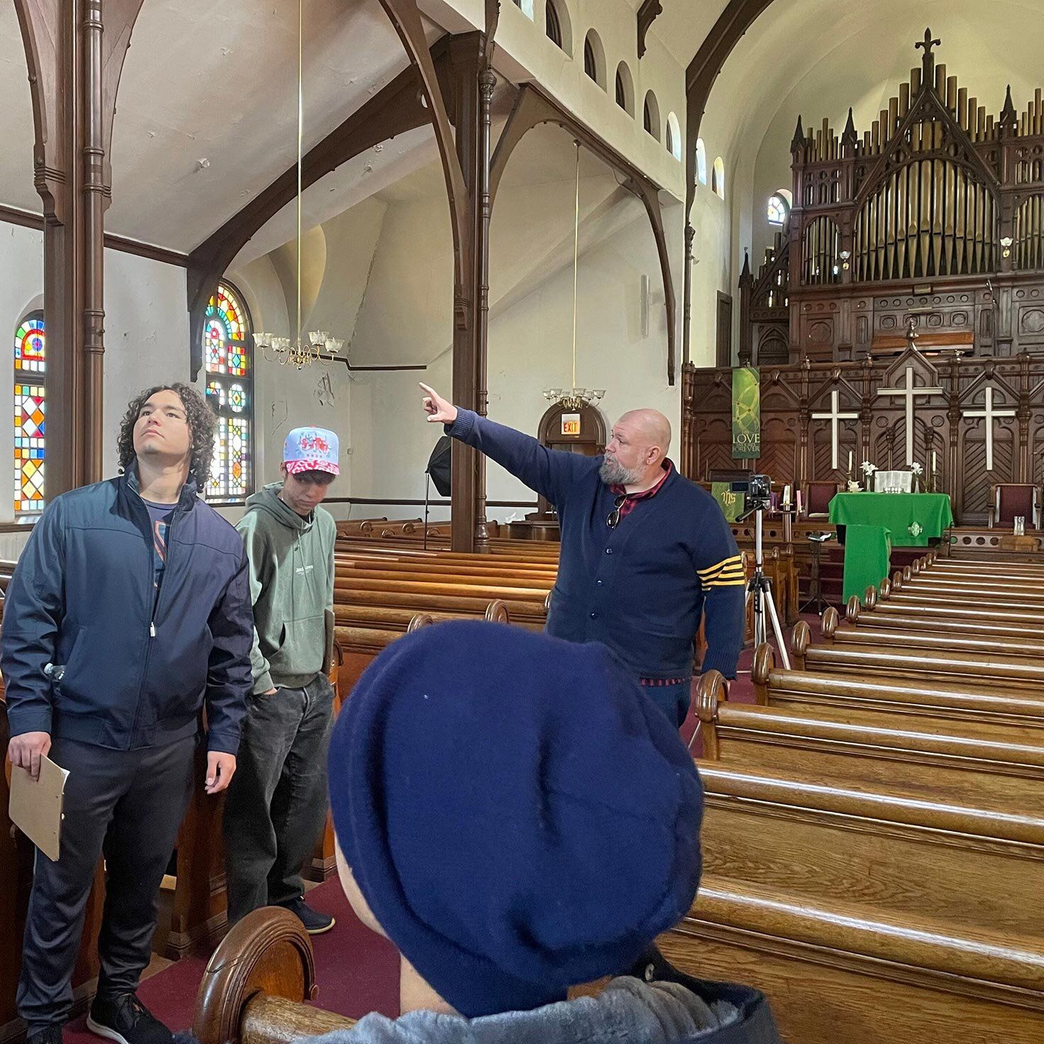 Visitors on an architecture tour explore a historic church interior featuring stained-glass windows, wood trusses, a pipe organ and rows of wooden pews.
