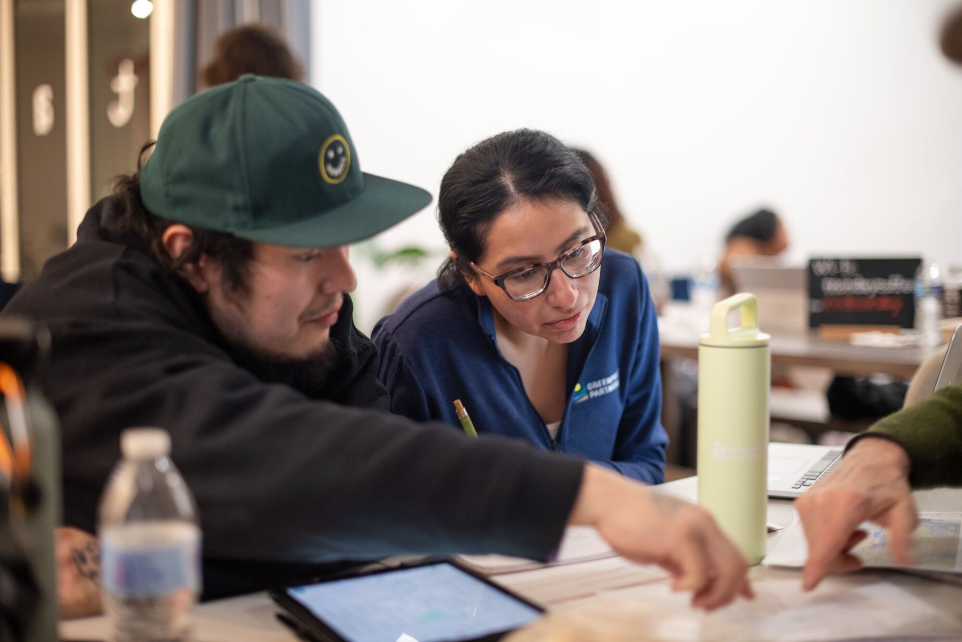 Man in a green hat and woman with glasses in a blue button up looking down at papers on a table. 