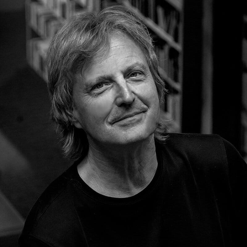 Black-and-white portrait of architect Tom Kundig seated indoors, looking toward the camera, with bookshelves softly blurred in the background
