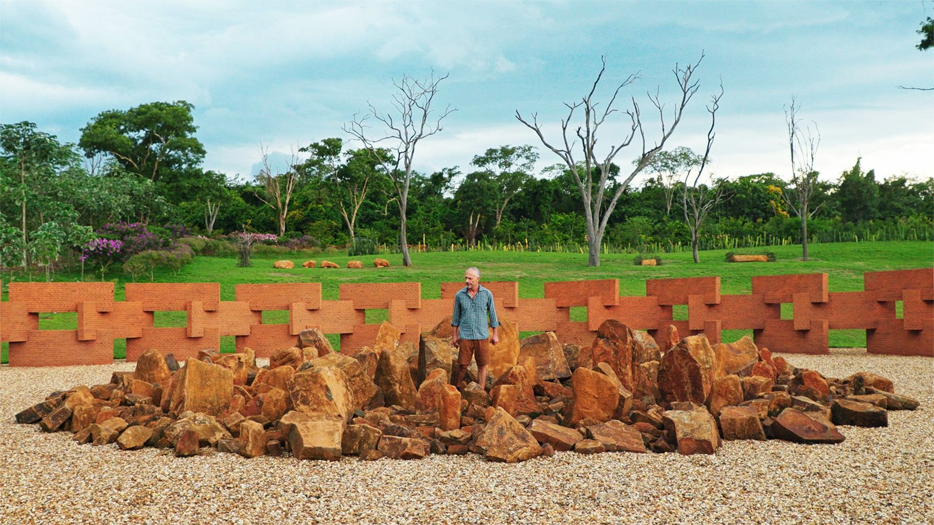 One man standing in a midst of a pile or red rocks with a brick sculpture in the background