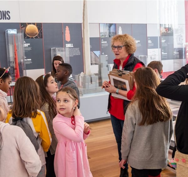 A group of children in a gallery overseen by a person with short hair in red shirt and black vest