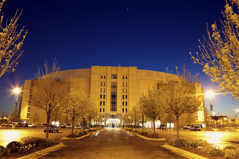 Symmetrical view of a large arena with a curved roof, seen from a central driveway lined with flowering trees and illuminated by parking lot lights at dusk.
