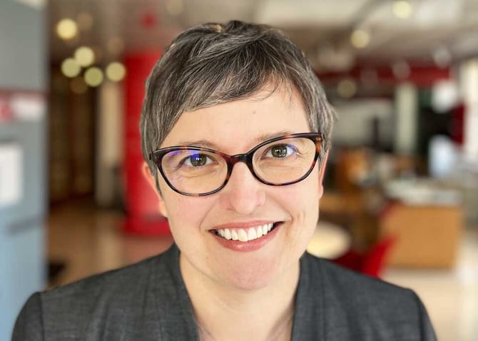 Portrait of a person wearing glasses and a gray blazer, photographed indoors with a softly blurred background.