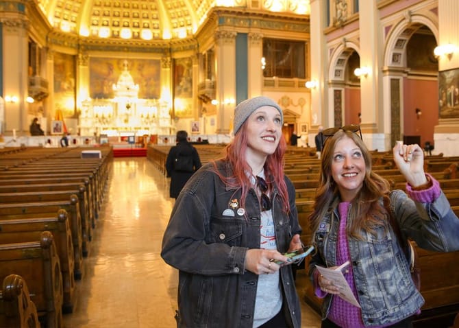 Two women with long hair standing in the foreground pointing and looking up while smiling with a church setting in the background. 