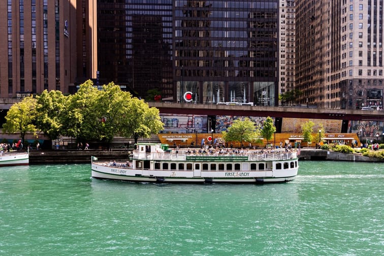 The top of a packed river cruise boat going down the river with skyscrapers surrounding river banks.