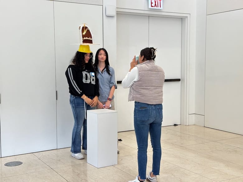 Two teens, one wearing a handmade headpiece pose for a photo taken by a woman standing in front of them. 