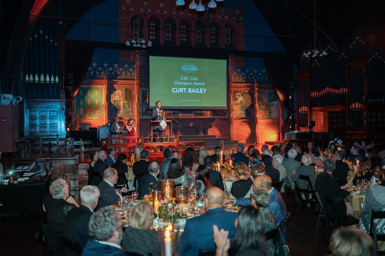 A full event with people seated at round tables and a stage in front of them with a speaker standing in front of a screen that reads "CAC Civic Champion Award Curt Bailey