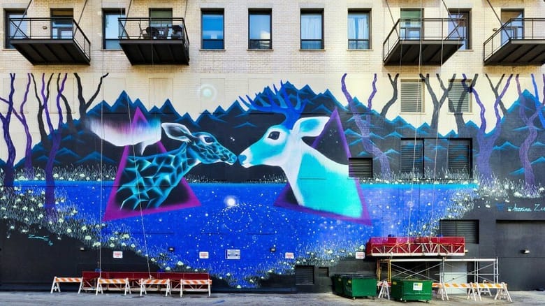 Large mural of two deer on blue background on side of building with scaffolding.