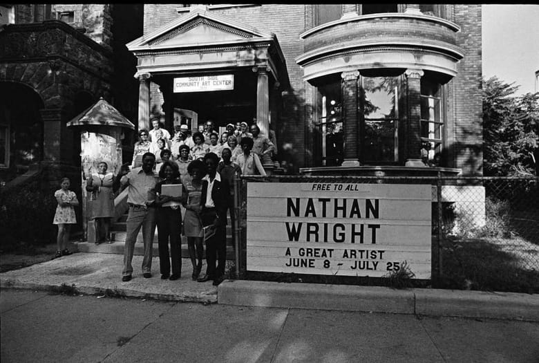 Black and white, exterior shot of building with people standing outside. Sign outside says Nathan White, free for all