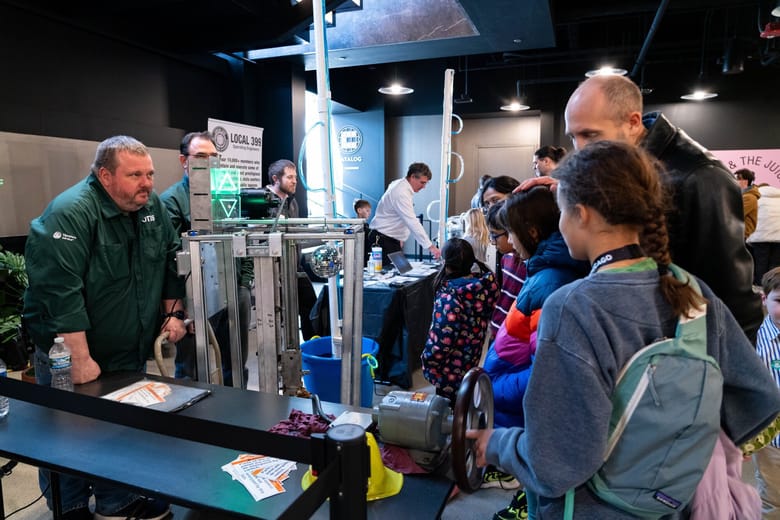 A man in a green zip up jacket stands across from a group of girls and a man with engineering equipment between them.