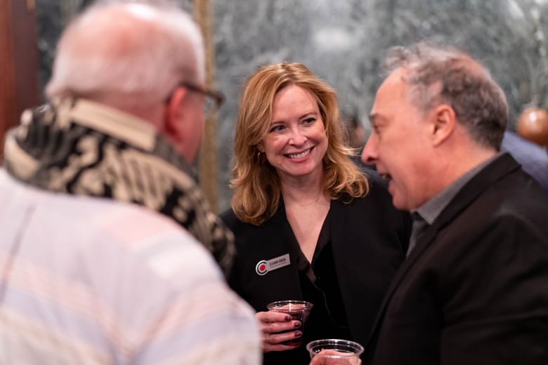 Image of a woman smiling with a man to her right speaking and another man out of focus in the foreground.