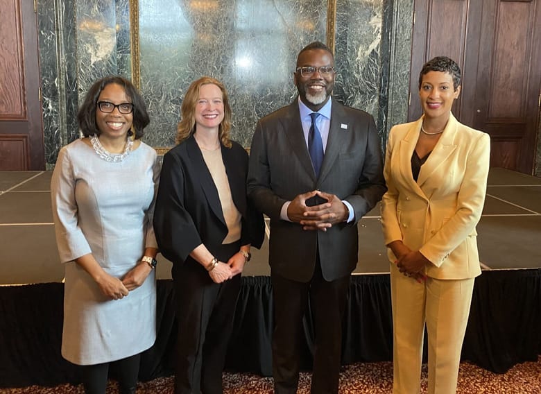 Four people standing shoulder to shoulder smiling and wearing business attire.