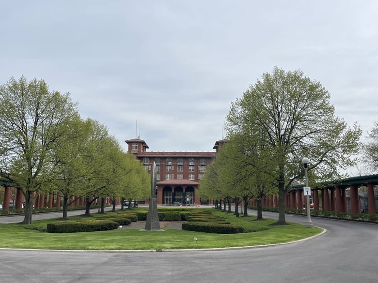 Large red brick building in the background and two lines of trees in the foreground. 