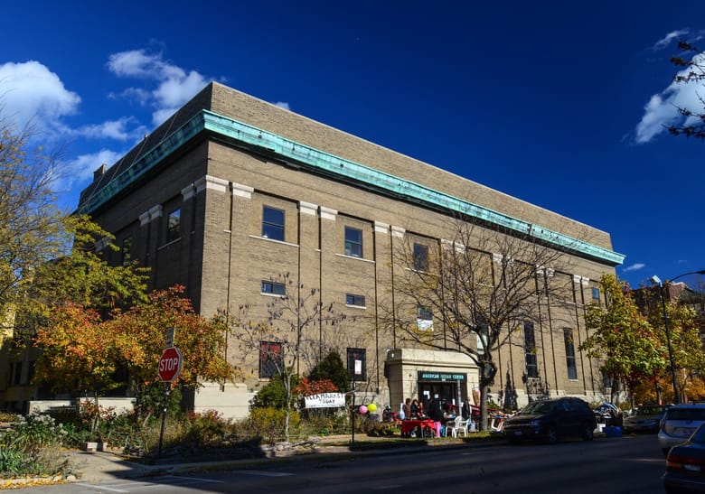 Exerior of a brown brick building with a green stripe around the top and trees outside.