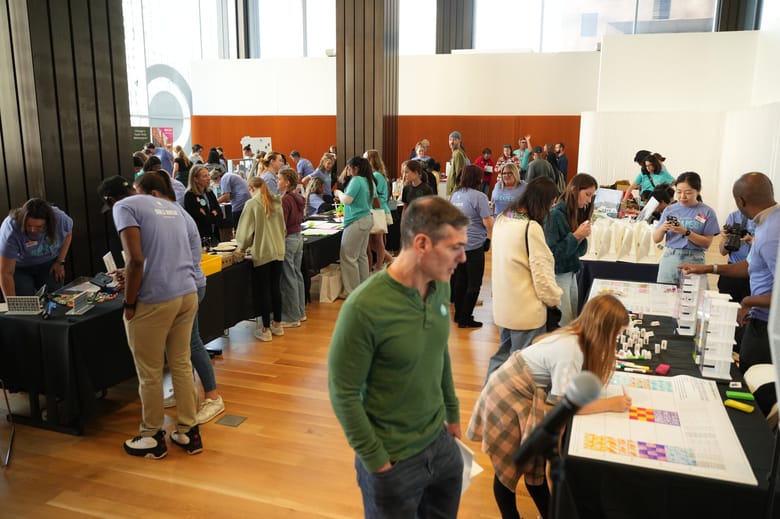 A crowd of people in a large room gather around lines of tables on each side.