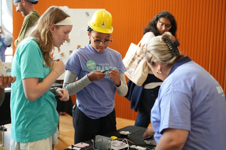 Two girls stand in front of a table with wiring on it looking at materials. A woman stands facing away on the other side of the table in the foreground.