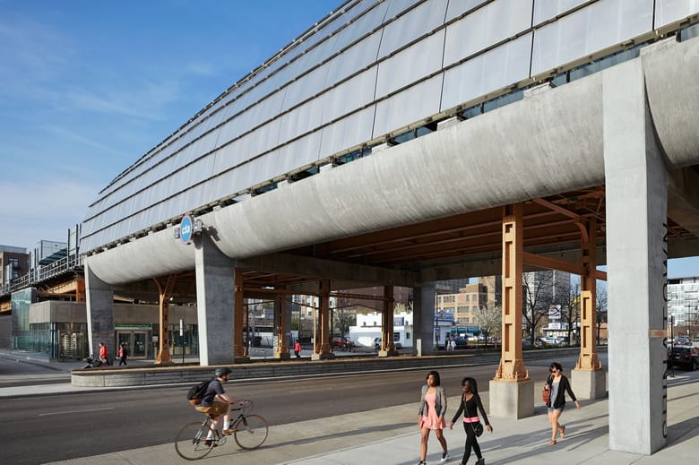 Image of an overpass structure with large metal shingles on the side and people waling below. 