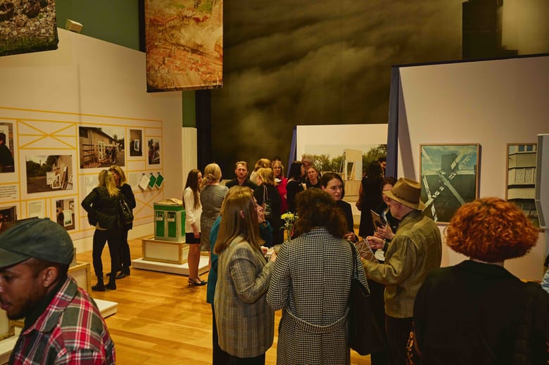 Image of a gathering of several people in conversation standing in a large exhibition space with images on the wall and models in the room.
