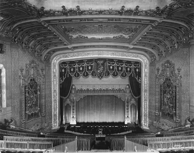 black and white image of the interior of a large theater looking at the stage from a balcony. 