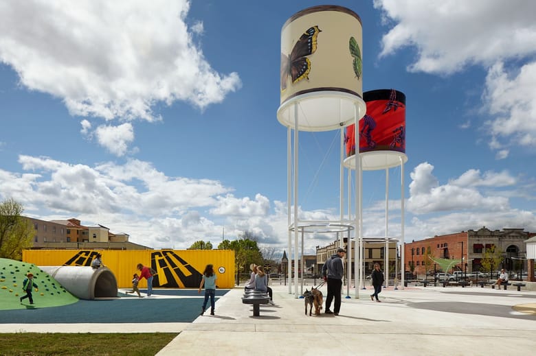 Two structures resembling water towers with colorful paintings on the side and people standing in a park below with brightly colored structures. 