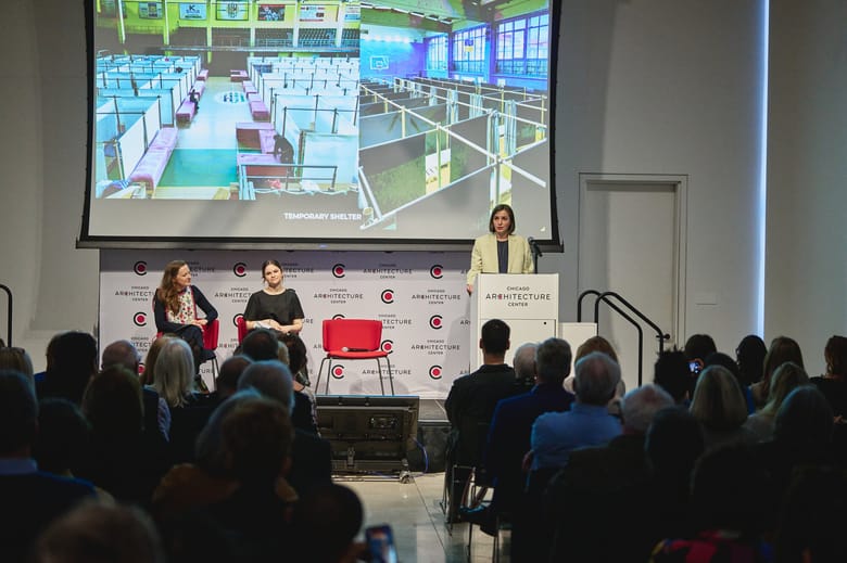 Three women on a stage with a projector screen behind them. One is standing behind a podium on the right and the other two are seated in chairs to the left. 