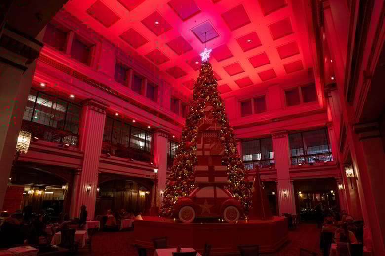 large room lit in red lights with high ceilings and a giant Christmas tree with s large toy car in front of it as decor.