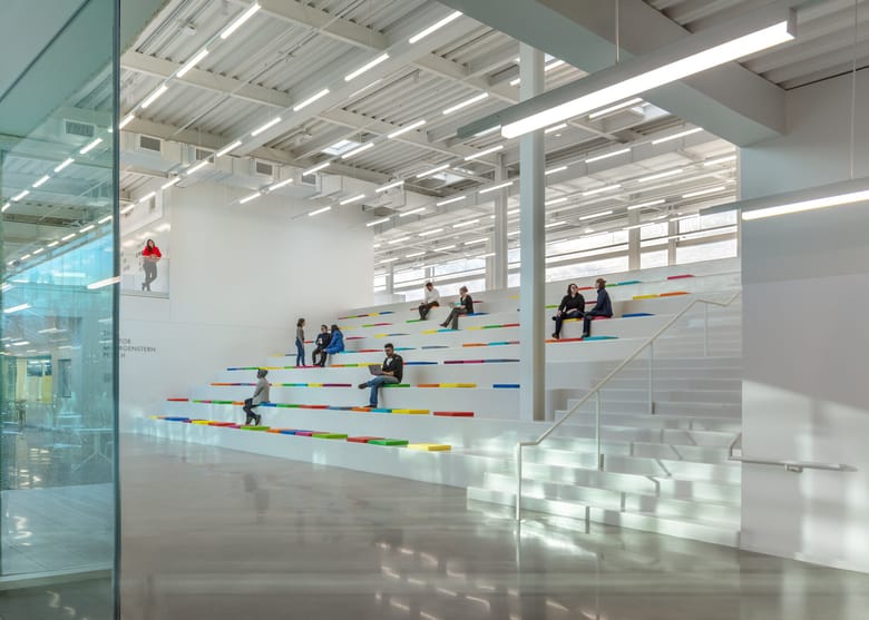 Large white room with tall ceilings and steps that have colorful cushions and people sitting at various levels working.