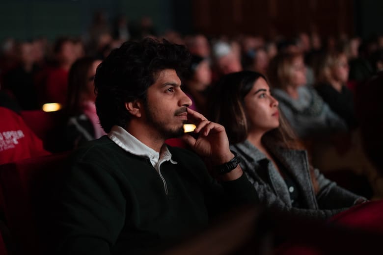 Close up image of two people sitting in a theater looking at a screen that is out of frame with other audience members in the background. 