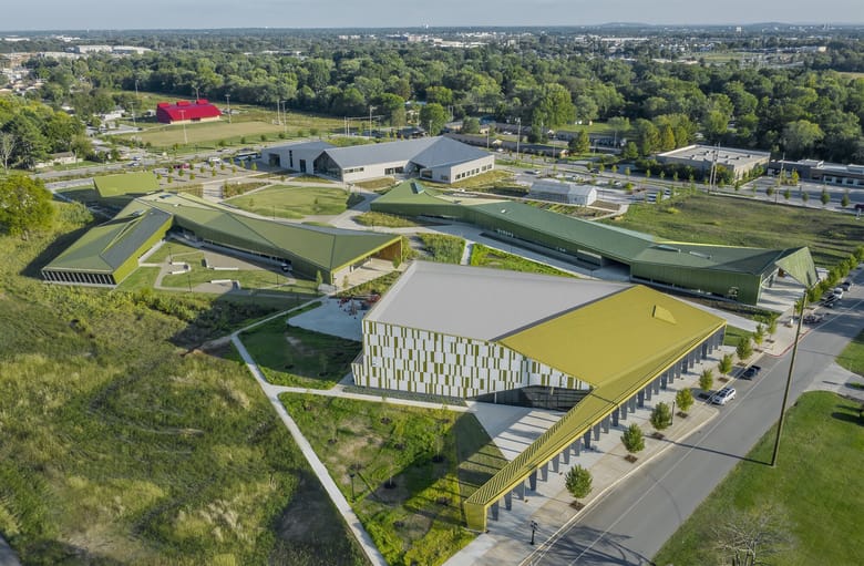 View from above looking down on a green field with trees in the background and a large building in the foreground. 
