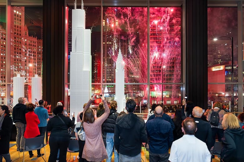 People inside a gallery looking outside the building where there are red and pink fireworks.