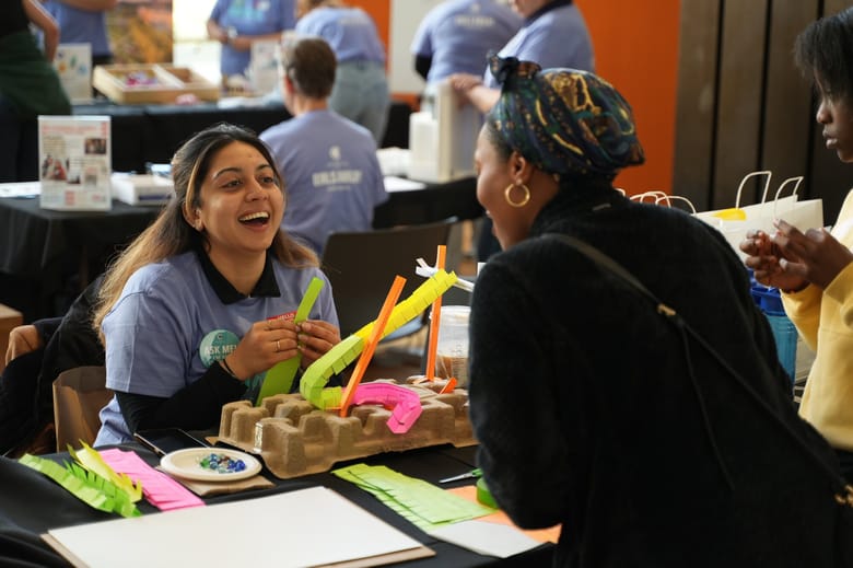 Two women sitting at a table with craft supplies one facing the camera and smiling the other faced away. 