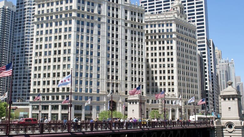 Looking across bridge and river at large white building. Several pedestrians and flags on bridge. 