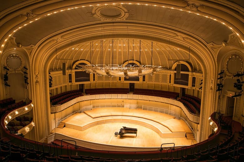 Interior of a concert hall view from the balcony looking down at an empty lit stage with a piano in the center. 