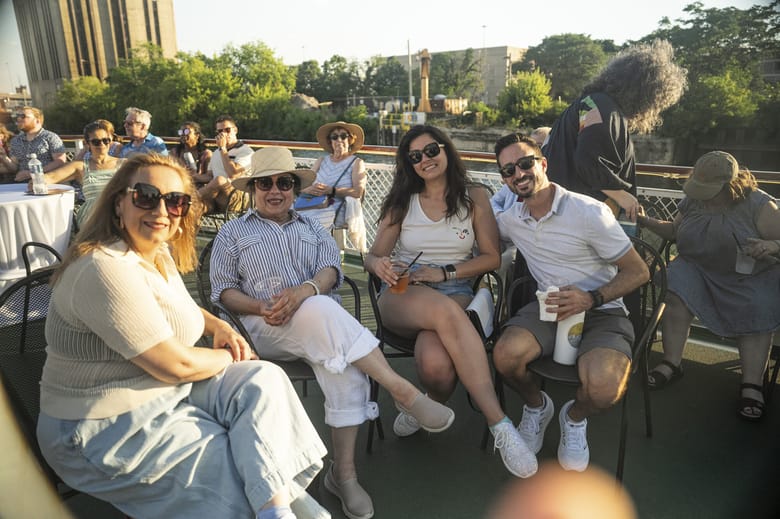 Four people in the foreground wearing sunglasses and smiling while sitting on a boat deck with other people in the background. 