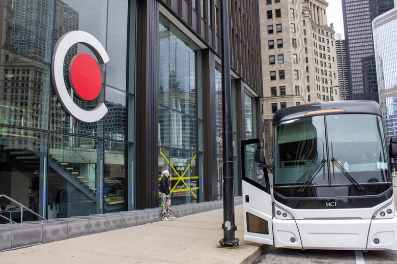 The face of a tour bus parked next to a black building with tall windows.