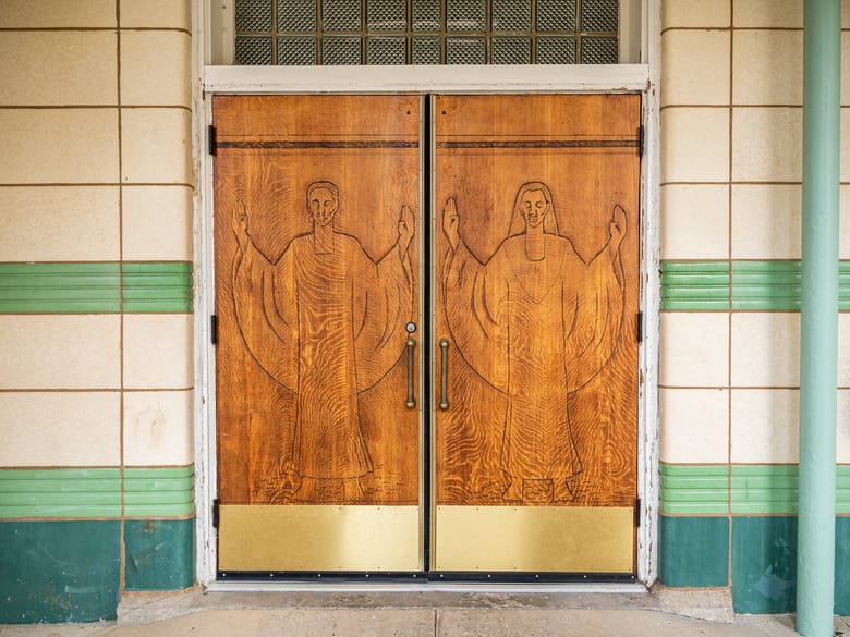 Image of large wooden double doors with relief carvings of a man and woman with their arms raised.